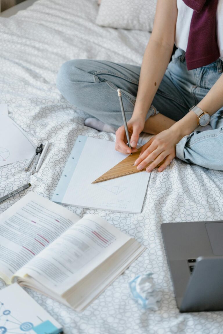 Student using set square and textbooks while studying on a bed at home.